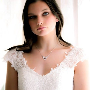 May include: A woman wearing a white lace wedding dress with a delicate silver necklace featuring a floral design.