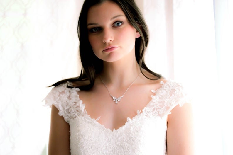 May include: A woman wearing a white lace wedding dress with a delicate silver necklace featuring a floral design.