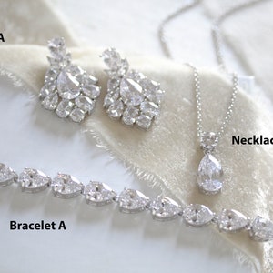 May include: A bridal jewelry set with a silver necklace, bracelet, and earrings. The necklace features a teardrop-shaped pendant with clear stones. The bracelet and earrings are also adorned with clear stones.