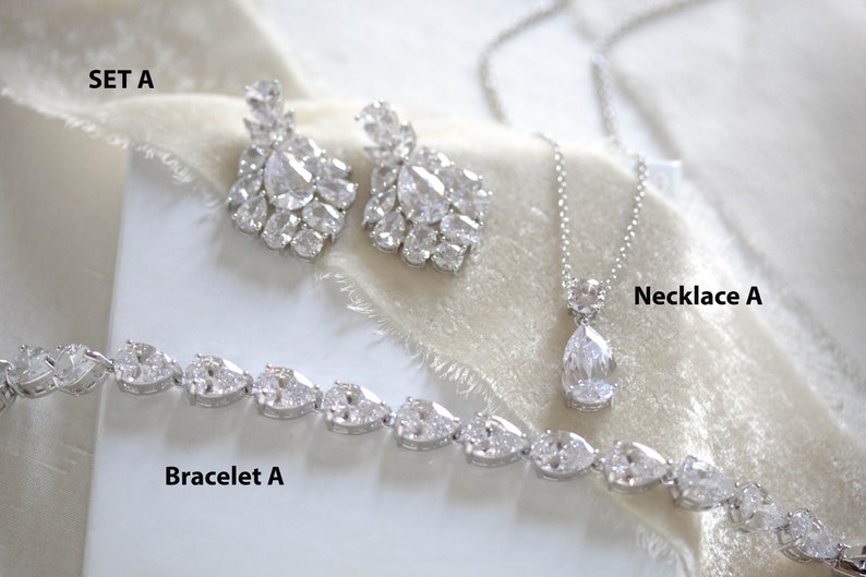 May include: A bridal jewelry set with a silver necklace, bracelet, and earrings. The necklace features a teardrop-shaped pendant with clear stones. The bracelet and earrings are also adorned with clear stones.