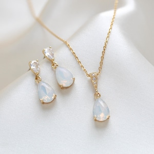Op de afbeelding: Een goudkleurige sieradenset met een ketting en bijpassende oorbellen. De ketting heeft een fijne ketting met een hanger en de oorbellen zijn in druppelstijl. Elk stuk heeft een peervormige, opalescerende steen en heldere accentstenen.