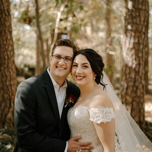 May include: A bride and groom pose for a wedding portrait in a forest setting. The bride is wearing a white lace dress with a veil and the groom is wearing a black suit. They are smiling and looking at the camera.