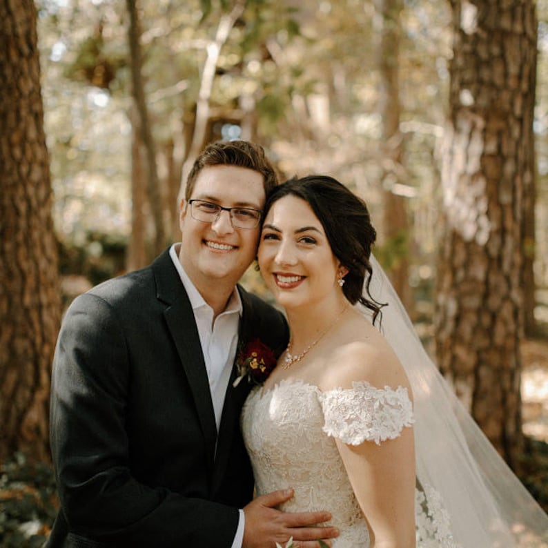 May include: A bride and groom pose for a wedding portrait in a forest setting. The bride is wearing a white lace dress with a veil and the groom is wearing a black suit. They are smiling and looking at the camera.
