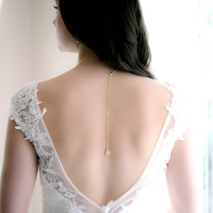 May include: A woman wearing a white lace dress with a low back, showcasing a delicate gold chain with a single teardrop-shaped pendant.