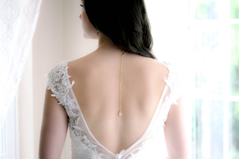 May include: A woman wearing a white lace dress with a low back, showcasing a delicate gold chain with a single teardrop-shaped pendant.