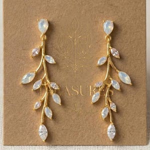 May include: A pair of gold-toned drop earrings featuring a branch-like design adorned with marquise-cut iridescent white stones. The earrings are attached to a rectangular brown card with gold foil text that reads TREASURES.