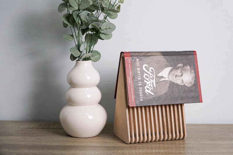 May include: A wooden book stand with a book titled "Henry Ford, My Life and Work" resting on top. The book stand is made of light wood with a series of vertical slats. A white vase with a bulbous shape holds a sprig of green foliage.