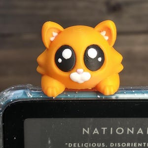 May include: An orange cat-shaped bookmark with large black eyes and a white nose. The bookmark is resting on a book with the word "NATIONAL" visible. The book also has the text "DELICIOUS, DISORIENTING" and the author's name.