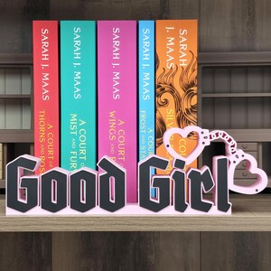 May include: A decorative sign featuring the text Good Girl in a bold black font with a light pink base. The design includes a light pink heart-shaped handcuff motif attached to the letter l, positioned in front of five colorful book spines.