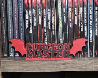 Może przedstawiać: Trójwymiarowy znak w kolorze czerwonym i czarnym z napisem "DUNGEON MASTER" pogrubioną czcionką, otoczony z obu stron stylizowanymi skrzydłami nietoperza. Znak umieszczony jest na półce przed rzędem różnych książek do gier fabularnych.