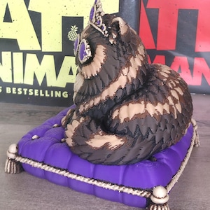 Peut inclure: Figurine de hibou d&eacute;taill&eacute;e avec une couronne, pos&eacute;e sur un coussin violet avec une bordure en corde blanche et des glands. Le hibou a des plumes marron, beige et noires et des accents violets. En arri&egrave;re-plan, des couvertures de livres.
