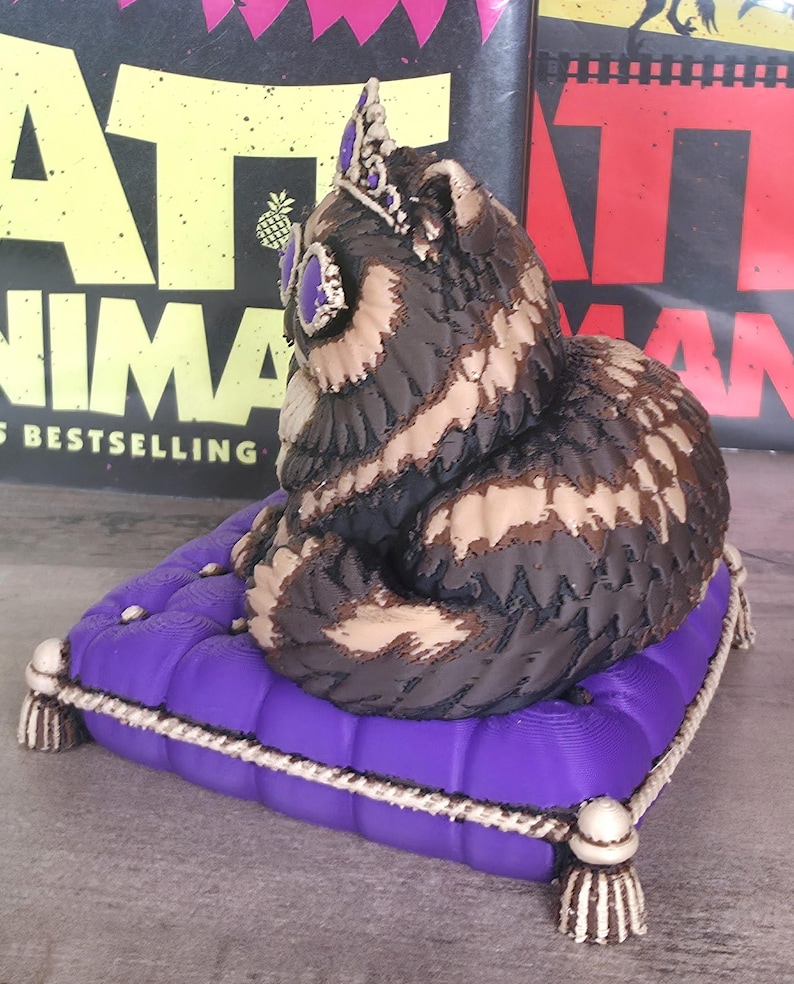 Peut inclure: Figurine de hibou d&eacute;taill&eacute;e avec une couronne, pos&eacute;e sur un coussin violet avec une bordure en corde blanche et des glands. Le hibou a des plumes marron, beige et noires et des accents violets. En arri&egrave;re-plan, des couvertures de livres.