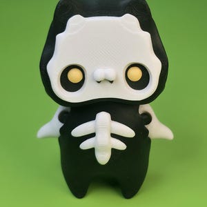 May include: A small, stylized figurine featuring a black and white color scheme. The character has a large white face mask with black eye sockets containing yellow circular pupils, a black body, and a white skeletal rib cage design on the chest.