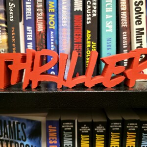 May include: A bright orange 'THRILLER' word art piece is displayed in front of a collection of books. The bold lettering is a decorative accent for a bookshelf or desk, ideal for book enthusiasts.