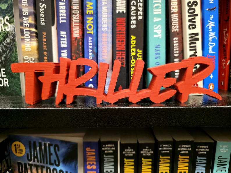 May include: A bright orange 'THRILLER' word art piece is displayed in front of a collection of books. The bold lettering is a decorative accent for a bookshelf or desk, ideal for book enthusiasts.