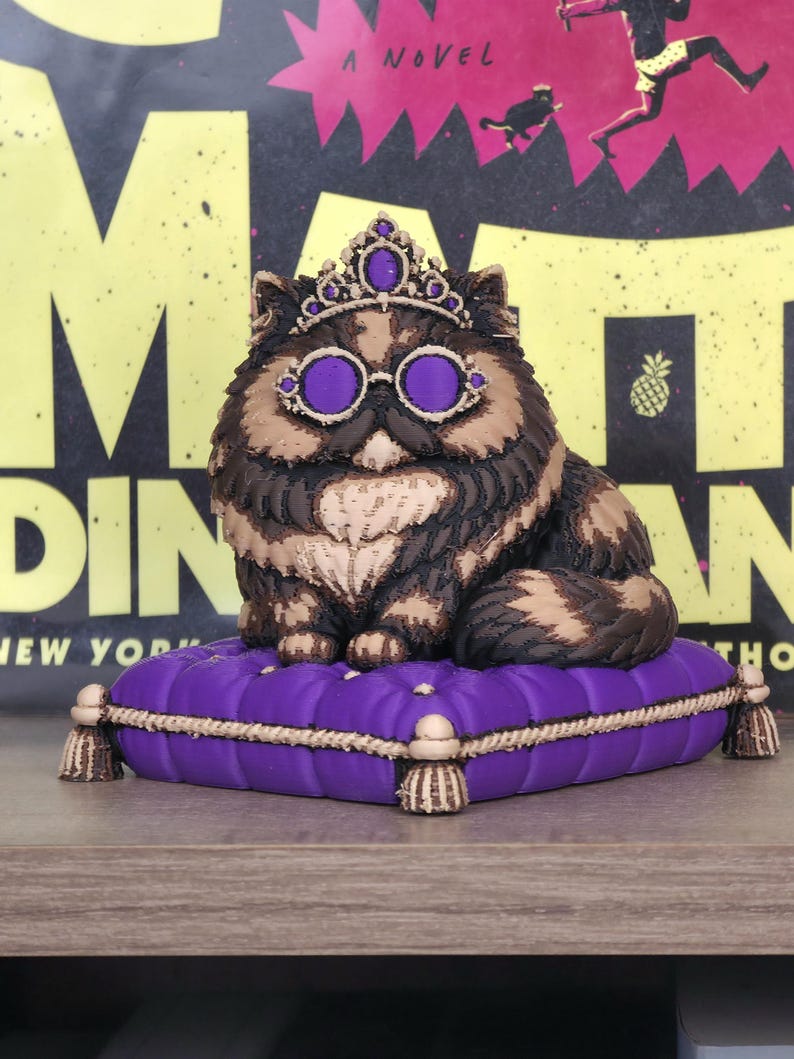 Peut inclure: Figurine de chat royal avec une couronne et des lunettes violettes, pos&eacute;e sur un coussin violet. Le chat a un pelage brun et beige. Le coussin est orn&eacute; de bordures et de glands dor&eacute;s. En arri&egrave;re-plan, une couverture de livre.