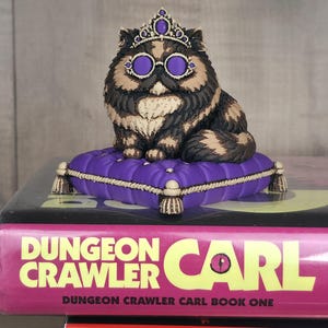 Peut inclure: Figurine de chat orn&eacute;e d'une couronne et de lunettes violettes, assise sur un coussin violet avec des bordures dor&eacute;es. Le chat est brun, noir et cr&egrave;me. La figurine est pos&eacute;e sur un livre intitul&eacute; "Dungeon Crawler Carl".