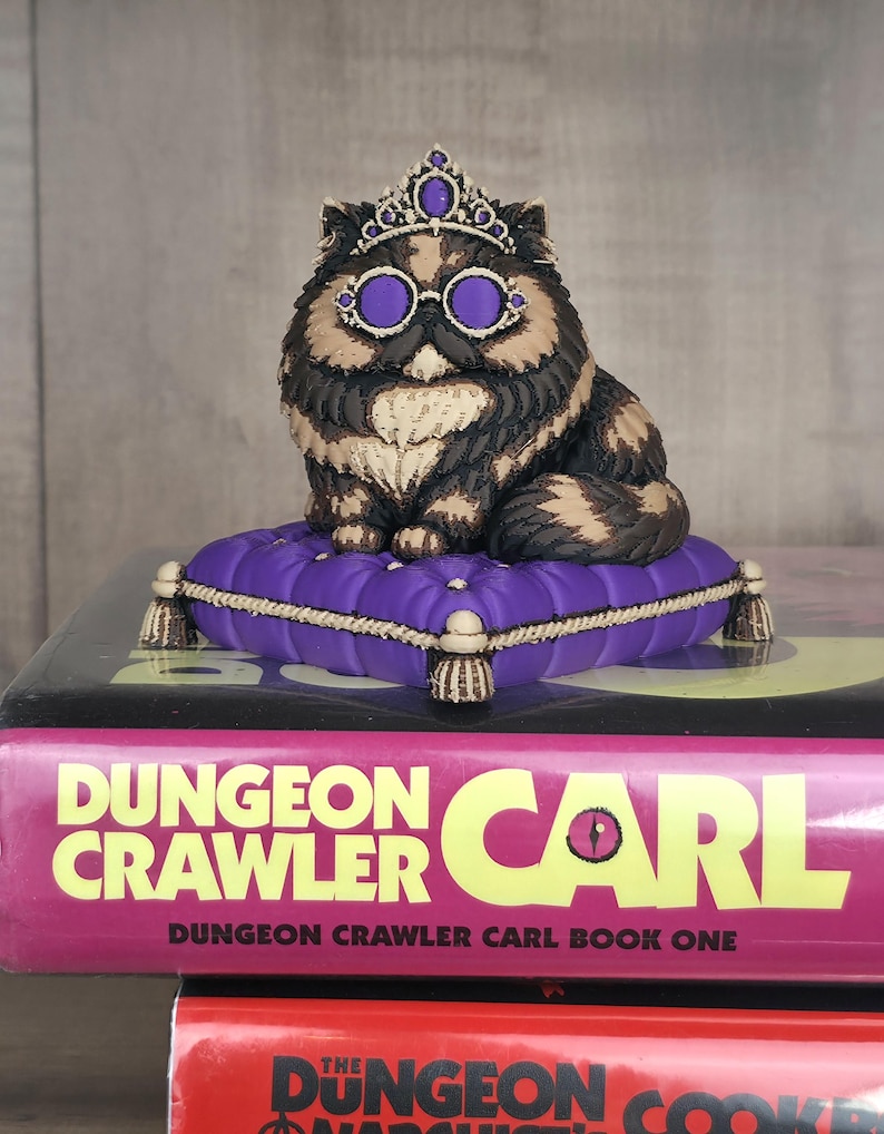 Peut inclure: Figurine de chat orn&eacute;e d'une couronne et de lunettes violettes, assise sur un coussin violet avec des bordures dor&eacute;es. Le chat est brun, noir et cr&egrave;me. La figurine est pos&eacute;e sur un livre intitul&eacute; "Dungeon Crawler Carl".