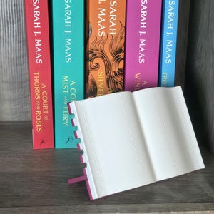 May include: A collection of colorful books by Sarah J. Maas, with titles including "A Court of Thorns and Roses" and "A Mist and Fury." The books are arranged vertically, with an open book in the foreground.
