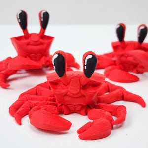 May include: Three bright red crab figurines with articulated legs and claws. Each crab features two prominent, upright eye stalks with black, oval-shaped eyes that have a small white highlight. The surface of the crabs shows a layered texture.