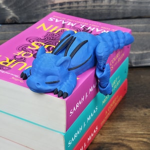 May include: A blue dragon bookend rests atop a stack of books with pink, teal, and red covers. The dragon has black accents and is lying down. The books are by Sarah J. Maas.