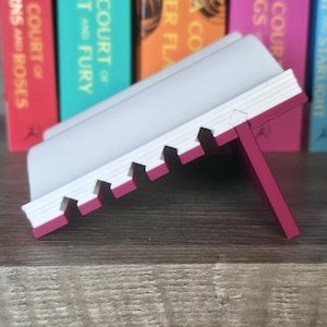 May include: A white and pink book stand with a notched design. The stand is angled, with a pink support leg. The background features a row of colorful books with titles like "A Court of Thorns and Roses" and "A Court of Mist and Fury."