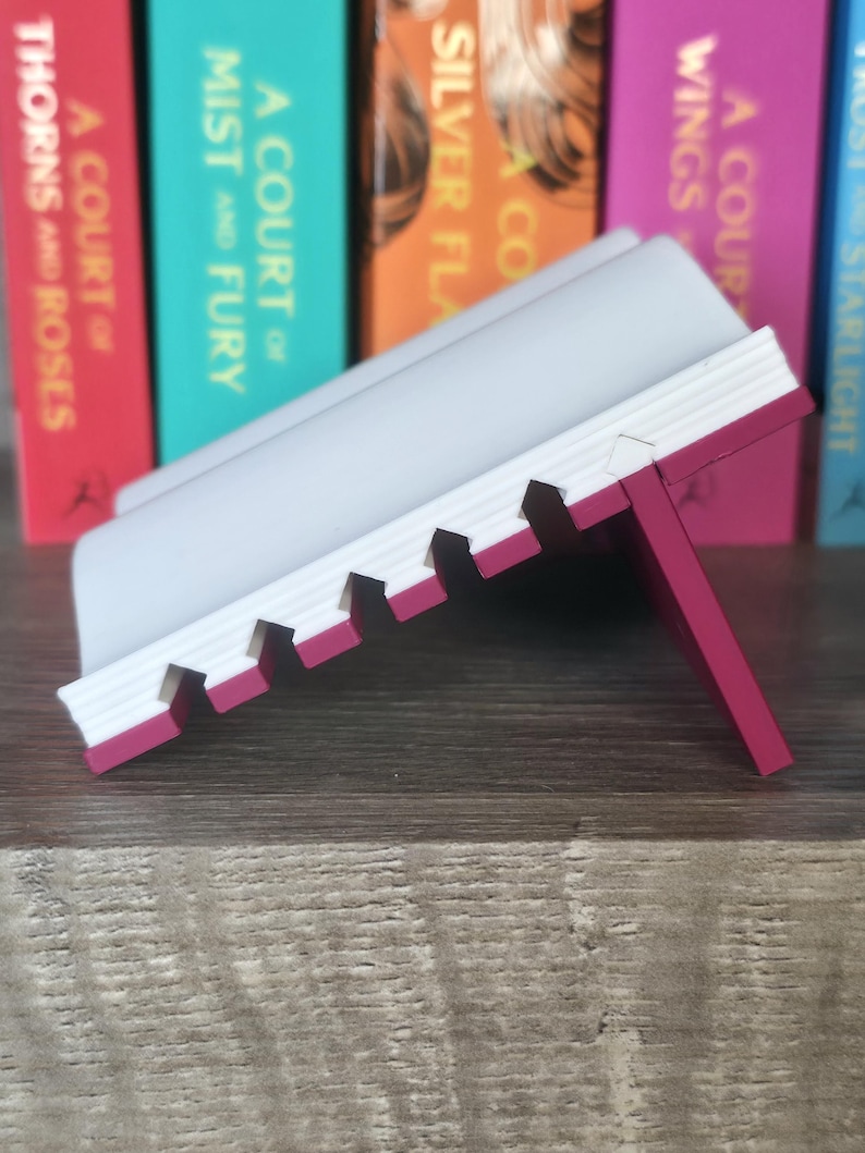 May include: A white and pink book stand with a notched design. The stand is angled, with a pink support leg. The background features a row of colorful books with titles like "A Court of Thorns and Roses" and "A Court of Mist and Fury."
