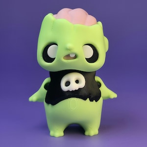 May include: A green zombie figurine with a pink brain exposed. The figure has large black eyes, a small mouth, and a black shirt with a white skull design. The background is a solid purple.