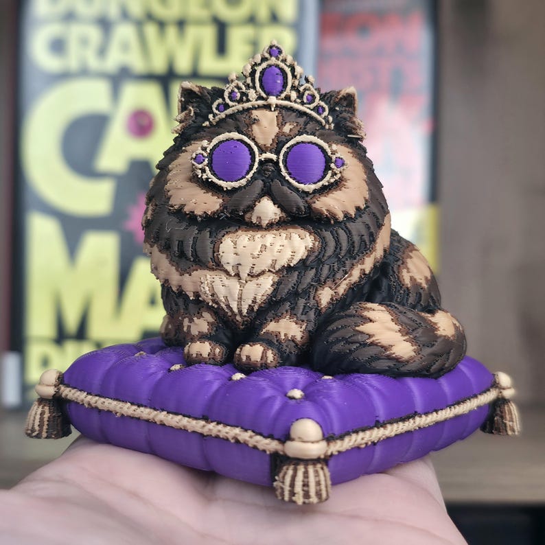 Peut inclure: Figurine d&eacute;taill&eacute;e d'un chat portant une couronne et des lunettes teint&eacute;es de violet, reposant sur un coussin violet avec une bordure dor&eacute;e. Le chat a des poils bruns, beiges et noirs. Le coussin a des glands d&eacute;coratifs &agrave; chaque coin. L'arri&egrave;re-plan est flou.