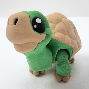 May include: A 3D-printed turtle figurine with a tan head and shell, and green body and limbs. The turtle has large black eyes with white highlights, and a detailed, textured surface.