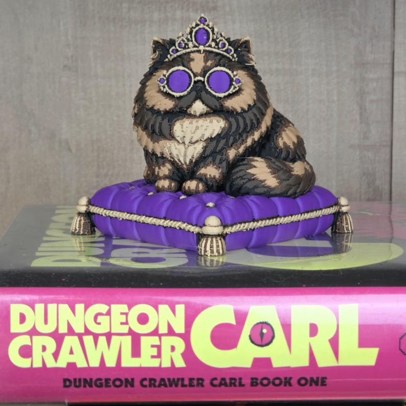 Peut inclure: Figurine de chat orn&eacute;e d'une couronne et de lunettes, assise sur un coussin violet avec des glands dor&eacute;s. Le chat a un pelage brun, noir et beige. Le coussin repose sur un livre intitul&eacute; "Dungeon Crawler Carl".