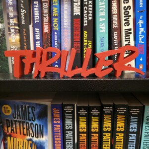 May include: A red "THRILLER" sign stands before a bookshelf packed with books. Titles include "Murder Island" and "The September House." Authors featured are James Patterson and Richard Osman. The books are in a home library.