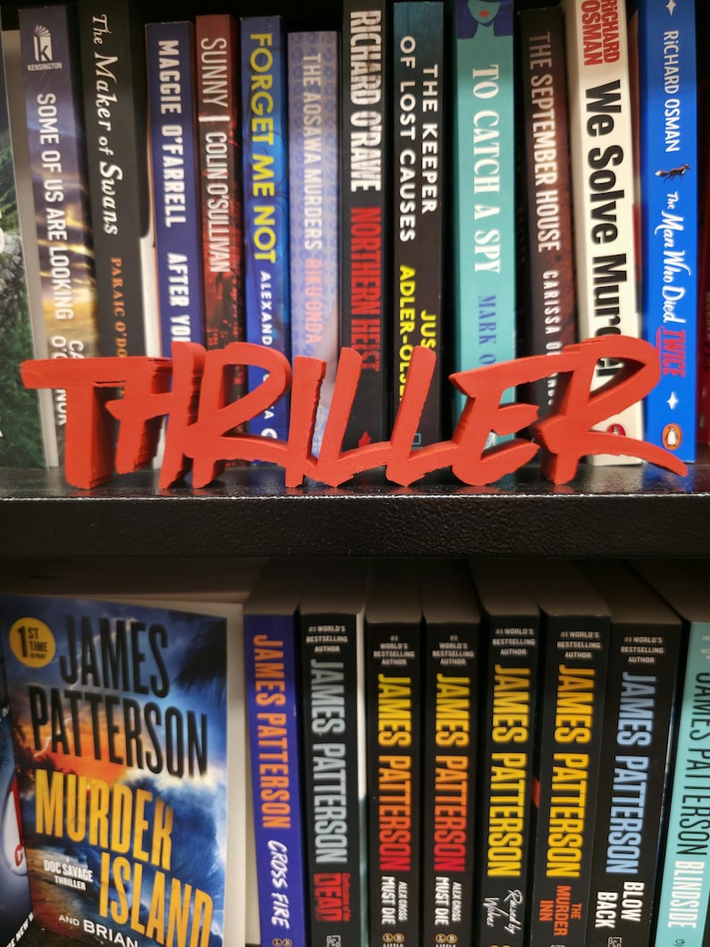 May include: A red "THRILLER" sign stands before a bookshelf packed with books. Titles include "Murder Island" and "The September House." Authors featured are James Patterson and Richard Osman. The books are in a home library.