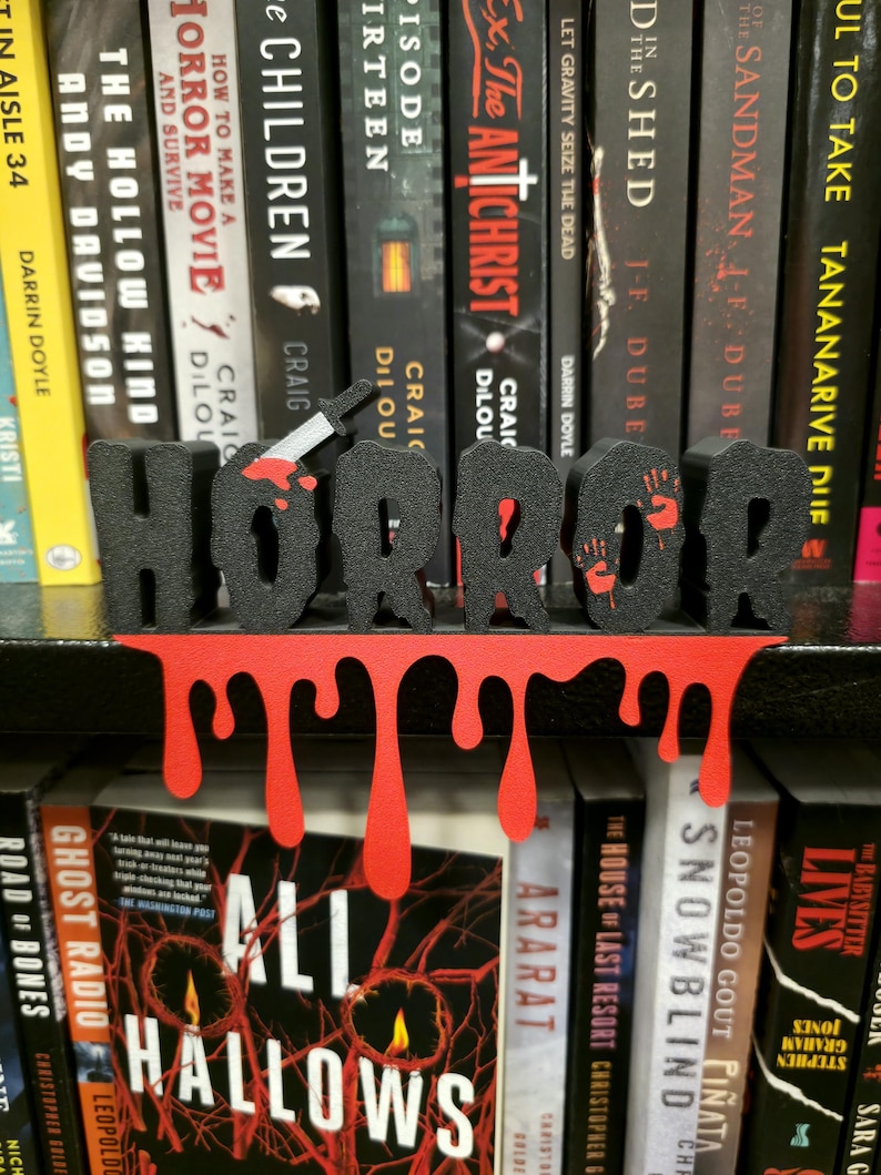 May include: A black "HORROR" bookend with a red blood drip design, displayed in front of a shelf of horror books. The bookend features a knife and handprints. The books have titles like "The Hollow Kind" and "All Hallows".