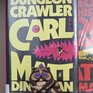 Peut inclure: Figurine de chat portant une couronne et des lunettes, assis sur un coussin violet, devant un livre intitul&eacute; "Dungeon Crawler Carl" de Matt Dinniman. La couverture du livre est noire avec du texte jaune et une illustration rose et rouge.