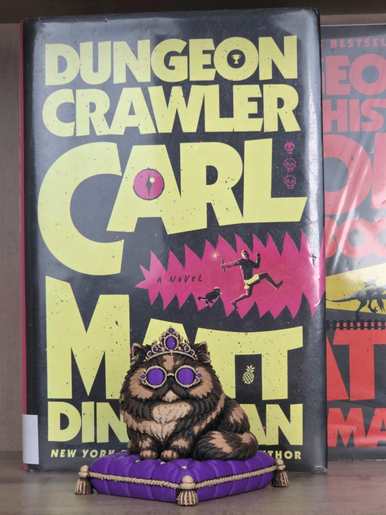 Peut inclure: Figurine de chat portant une couronne et des lunettes, assis sur un coussin violet, devant un livre intitul&eacute; "Dungeon Crawler Carl" de Matt Dinniman. La couverture du livre est noire avec du texte jaune et une illustration rose et rouge.