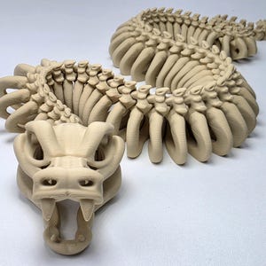 May include: A beige, articulated dragon sculpture with a detailed, skeletal design. The dragon's head features horns and sharp teeth, while its body is segmented for flexibility. The sculpture is made of a light-colored material and is displayed on a white surface.