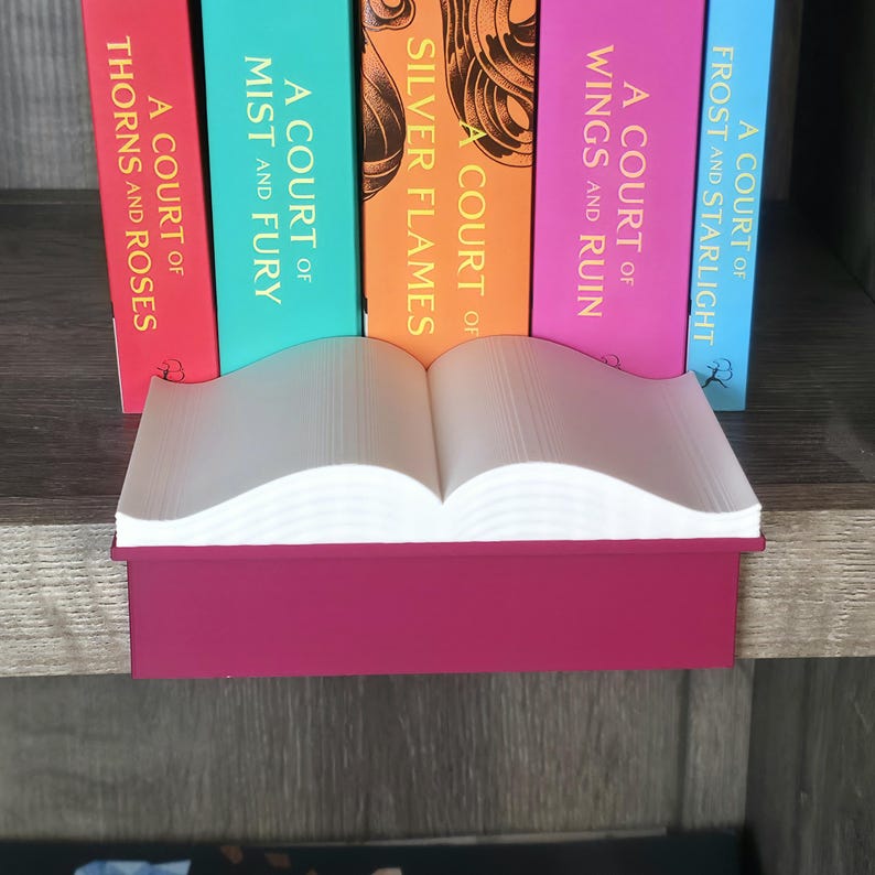May include: A burgundy-colored book-shaped shelf sitter with white pages, displayed in front of a row of colorful books. The books have titles like "A Court of Thorns and Roses" and "A Court of Mist and Fury."