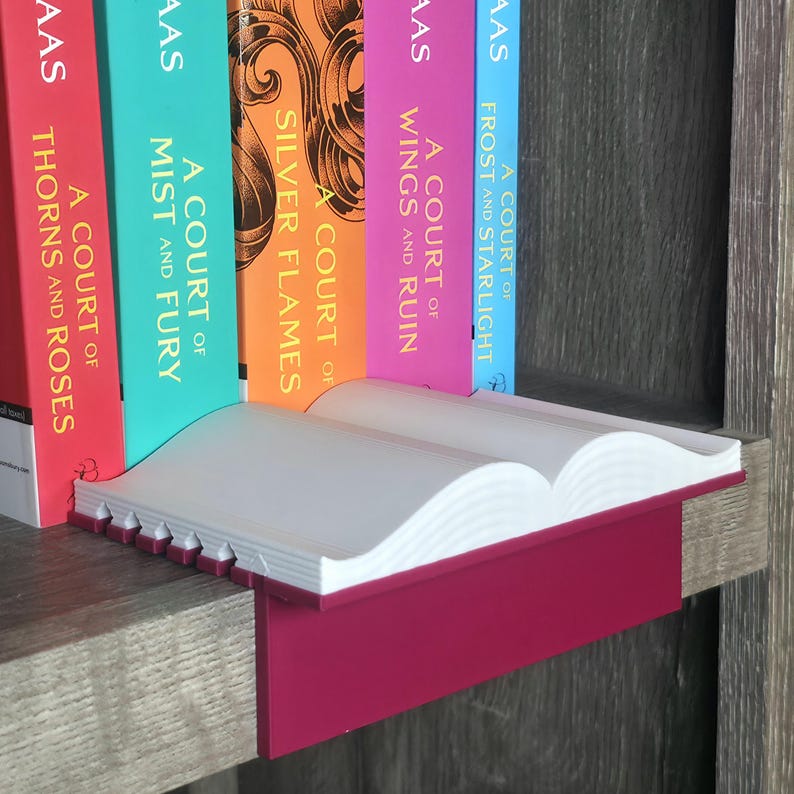 May include: A white and magenta book-shaped shelf with a row of colorful books. The books have titles like "A Court of Thorns and Roses" and "A Court of Mist and Fury". The shelf is on a wooden surface.