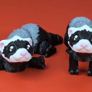 May include: Two 3D-printed ferret figurines. One is lying down, the other is standing. Both have black bodies, white faces with black markings, and gray backs. The ferrets have pink noses and are set against a red background.