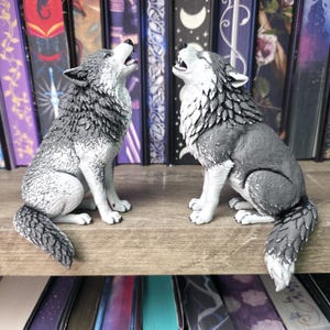 May include: Two gray and white wolf figurines in a sitting position with heads tilted upward as if howling. The wolves feature detailed fur textures and are positioned facing each other, showcasing their open mouths and sharp teeth.