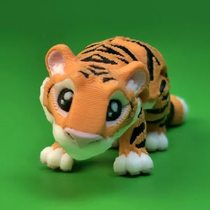 May include: A small 3D printed tiger figurine featuring orange fur with black stripes, large expressive eyes with white highlights, a light pink nose, and white paws. The textured surface shows visible layer lines from the printing process.