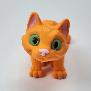 May include: Orange and pink 3D printed cat figurine with green eyes. The cat is sitting with its paws tucked under its body and looking directly at the camera.