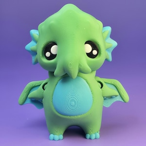 May include: A green and blue creature figurine with large, expressive eyes. The figurine has a rounded body, small wings, and a textured surface. The creature's belly and accents are a lighter shade of blue, set against a purple background.