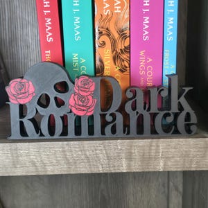 May include: A matte black 3D printed sign featuring the text "Dark Romance" in a serif font. The design includes a skull silhouette integrated into the letter D and two pink roses with black outlines positioned on the left side of the text.