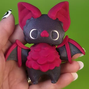 May include: A 3D-printed bat figurine with a dark gray body and wings, and vibrant magenta accents on the ears, chest, and wing edges. The bat has large, white crescent-shaped eyes and a small pink nose. The figurine is held in a hand against a green background.