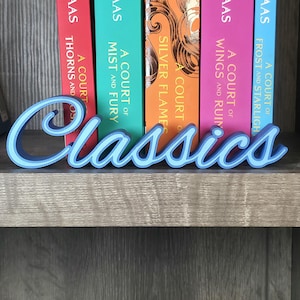 May include: A light blue "Classics" word sign in a cursive font, placed in front of a row of colorful books. The books have titles like "A Court of Thorns and Roses" and "A Court of Mist and Fury". The sign is on a wooden shelf.