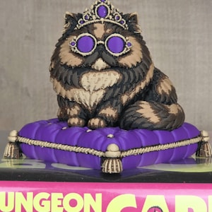 Peut inclure: Figurine de chat orn&eacute;e d'une couronne et de lunettes violettes, assise sur un coussin violet. Le chat a un pelage brun, noir et beige. Le coussin est bord&eacute; d'une corde dor&eacute;e et de glands.
