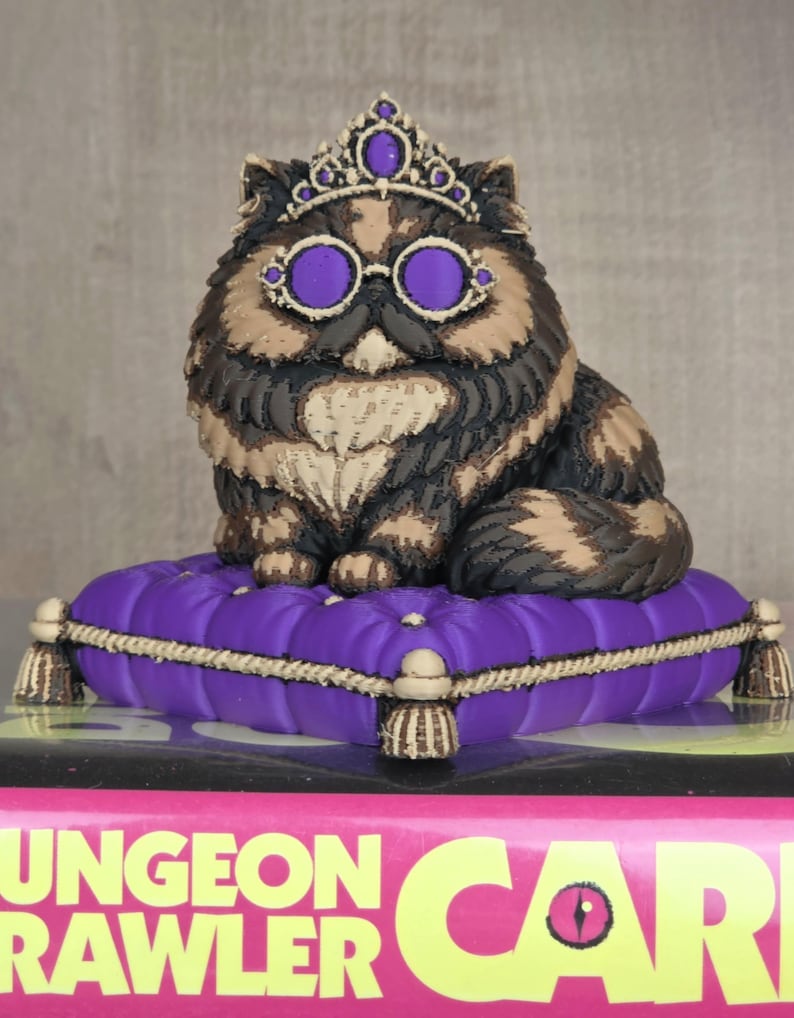 Peut inclure: Figurine de chat orn&eacute;e d'une couronne et de lunettes violettes, assise sur un coussin violet. Le chat a un pelage brun, noir et beige. Le coussin est bord&eacute; d'une corde dor&eacute;e et de glands.