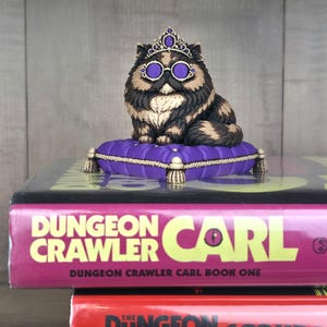 Peut inclure: Figurine d'un chat orn&eacute; d'une couronne et de lunettes, assis sur un coussin violet. Le chat est noir, marron et beige. Le coussin est pos&eacute; sur un livre intitul&eacute; "Dungeon Crawler Carl".